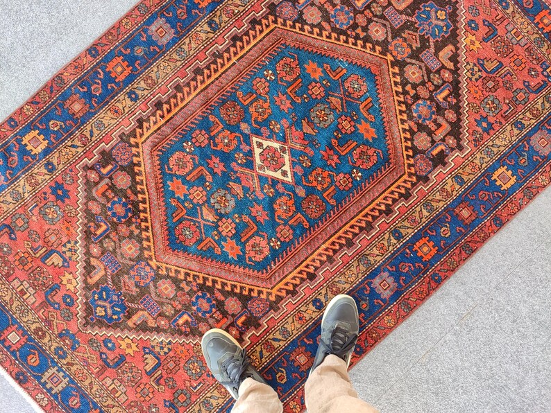 May include: A rectangular rug with a central blue geometric design, surrounded by a border of red, brown, and blue patterns. The rug features intricate floral and geometric motifs. The rug's dimensions are not visible in the image.