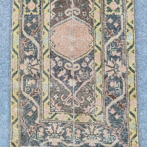 May include: A rectangular, vintage-style rug with a central diamond pattern in peach, surrounded by intricate floral and geometric designs. The rug features a color palette of muted greens, blues, yellows, and browns, creating a warm, inviting aesthetic. The rug's edges are slightly frayed, adding to its antique charm.