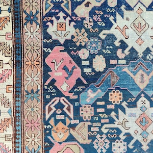 May include: A woven rug featuring a deep blue field with intricate geometric patterns in shades of pink, cream, and light blue. The design includes stylised animal figures and floral motifs, framed by a multi-layered border with repeating patterns.
