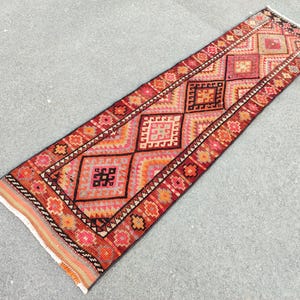 May include: A rectangular, hand-woven runner rug with a geometric pattern. The rug features a central design of repeating diamond shapes in shades of red, orange, and pink, bordered by a detailed floral pattern. The rug's fringe is visible at both ends.