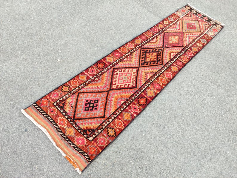 May include: A rectangular, hand-woven runner rug with a geometric pattern. The rug features a central design of repeating diamond shapes in shades of red, orange, and pink, bordered by a detailed floral pattern. The rug's fringe is visible at both ends.