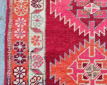 May include: Close-up of a patterned rug with a deep red backdrop. The design incorporates geometric shapes in pink, orange, and cream, with a border of floral patterns. The rug's texture is evident, indicating a woven construction.