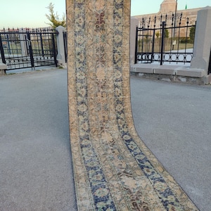 May include: A long, rectangular rug with an intricate, traditional design. The rug features a central beige field with a repeating pattern in shades of blue, red, and brown. The rug is displayed outdoors, partially rolled up.