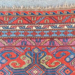 May include: Detailed view of a patterned rug with a traditional design. The rug displays a rich palette of red, blue, purple, and green, with intricate geometric and floral patterns. The edges have a fringed border.