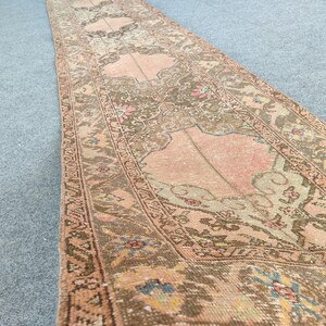 May include: A detailed runner rug with a floral design. The rug's central area is peach and brown, with green, blue, and pink accents. The border is a darker brown, and the rug is made of woven material. The rug is suitable for a hallway.