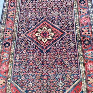 Peut inclure: Un tapis rectangulaire pr&eacute;sentant un champ bleu marine profond avec un motif g&eacute;om&eacute;trique r&eacute;p&eacute;titif complexe. Un m&eacute;daillon central en losange rouge corail contient un motif floral. La bordure affiche des &eacute;l&eacute;ments floraux rouges, bleus et cr&egrave;me.