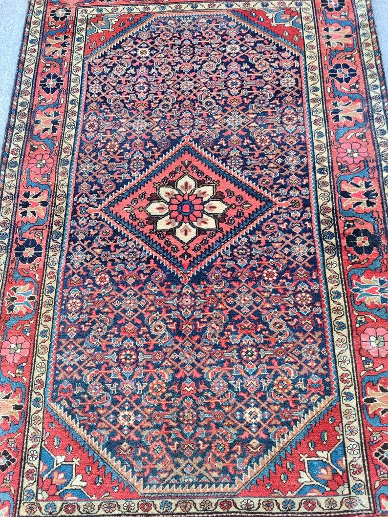 Peut inclure: Un tapis rectangulaire pr&eacute;sentant un champ bleu marine profond avec un motif g&eacute;om&eacute;trique r&eacute;p&eacute;titif complexe. Un m&eacute;daillon central en losange rouge corail contient un motif floral. La bordure affiche des &eacute;l&eacute;ments floraux rouges, bleus et cr&egrave;me.