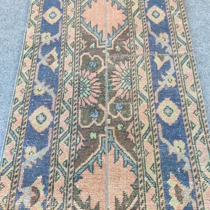 May include: A vintage rug with a distressed look. The rug has a central design in peach and brown, framed by blue and green borders. Geometric and floral patterns are visible throughout the rug's design.
