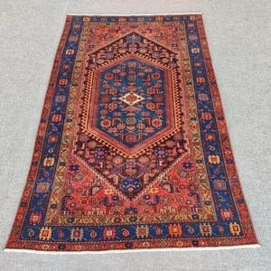 May include: A rectangular, hand-knotted rug with an intricate geometric design. The rug features a central medallion in shades of blue, brown, and red, surrounded by a border of blue and orange. The rug's dimensions are not visible in the image.