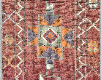 May include: Close-up of a hand-woven rug with a geometric pattern. The rug features a deep red background with orange, purple, and white geometric shapes. The design includes diamond and star-like motifs.