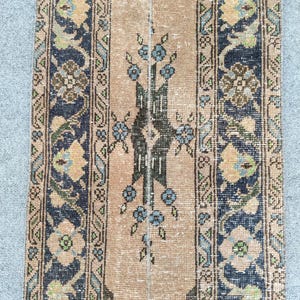 Könnte beinhalten: Ein rechteckiger Vintage-Teppich mit einem zentralen beigen Feld und aufwendigen Bordüren. Der Teppich weist florale und geometrische Muster in Marineblau, Beige und gedämpftem Grün auf. Das Design umfasst stilisierte Blumen und ein zentrales vertikales Motiv.
