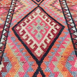 May include: Close-up of a colorful, patterned rug with a geometric design. The rug features a diamond pattern in shades of red, pink, orange, and black. The rug has a textured surface and a traditional style.