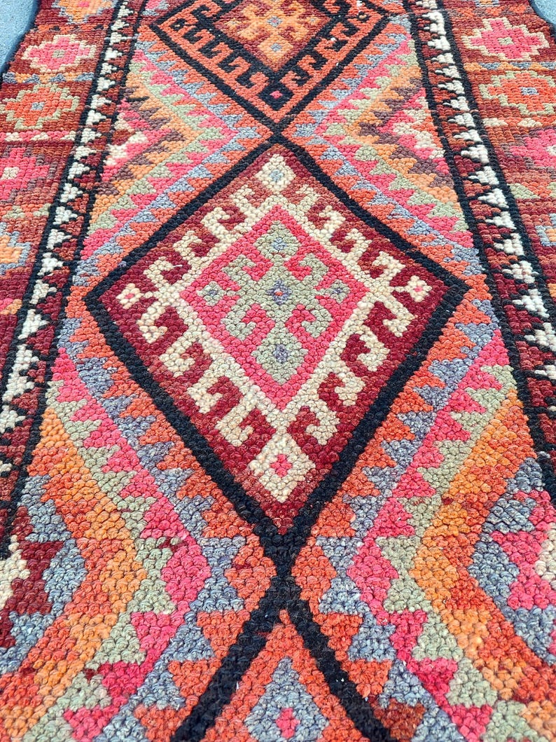 May include: Close-up of a colorful, patterned rug with a geometric design. The rug features a diamond pattern in shades of red, pink, orange, and black. The rug has a textured surface and a traditional style.