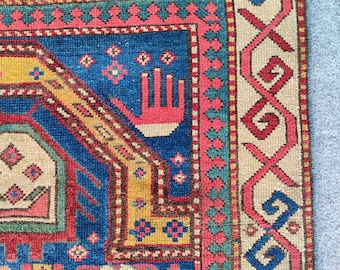 Op de afbeelding: Detail van een handgeweven tapijt met een complex geometrisch ontwerp. Het tapijt heeft een diepblauw middenveld met rode, gele en beige accenten. De rand heeft ingewikkelde patronen in rood en beige.