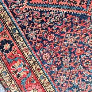 Peut inclure: Un tapis tiss&eacute; pr&eacute;sentant un champ bleu marine profond avec un motif dense et complexe de formes g&eacute;om&eacute;triques et de motifs floraux dans des tons de rouge corail, bleu clair et cr&egrave;me. La bordure affiche un motif floral r&eacute;p&eacute;t&eacute; sur fond rouge corail.
