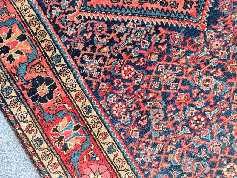 Peut inclure: Un tapis tiss&eacute; pr&eacute;sentant un champ bleu marine profond avec un motif dense et complexe de formes g&eacute;om&eacute;triques et de motifs floraux dans des tons de rouge corail, bleu clair et cr&egrave;me. La bordure affiche un motif floral r&eacute;p&eacute;t&eacute; sur fond rouge corail.