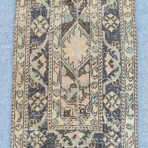 May include: A rectangular vintage rug with a central geometric design in beige, brown, and blue hues. The rug has a detailed border and a distressed look, indicating age and use. The pattern is symmetrical and intricate, typical of traditional rug designs.