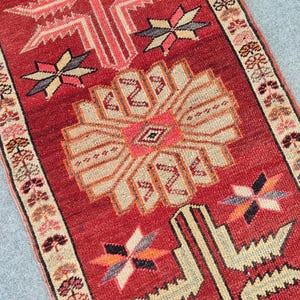 May include: A rectangular runner rug featuring a deep red background with a large central beige and pink geometric floral medallion. The rug is decorated with smaller star-shaped motifs and has a border with repeating floral patterns in beige, pink, and black.