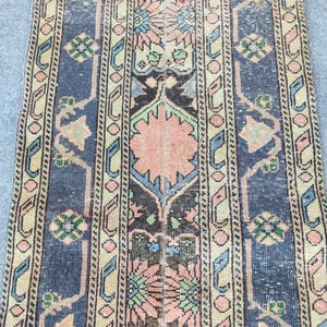 May include: A rectangular runner rug featuring a central vertical band with repeating floral motifs in peach, green, and dark brown, flanked by wide dark blue borders with geometric patterns and light tan stripes with small circular accents.