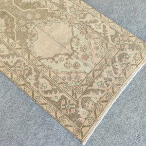 May include: A rectangular, vintage-style rug with a detailed, floral pattern. The rug features a beige central field with a darker border in shades of brown, green, and pink. The rug is placed on a grey surface.