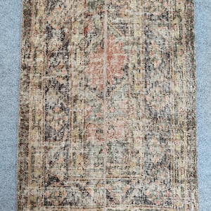 Könnte beinhalten: Ein rechteckiger, abgenutzter Teppich mit verblasstem Vintage-Aussehen. Der Teppich weist ein komplexes Muster in Braun-, Beige-, Grün- und Pfirsichtönen auf. Das Design umfasst florale und geometrische Elemente, die einen strukturierten, antiken Look erzeugen. Geeignet für verschiedene Innenräume.