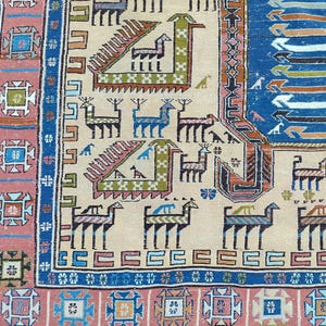 Puede incluir: Primer plano de una sección de una alfombra beige, azul y rosa estampada. La alfombra presenta diseños geométricos y figuras de animales estilizadas en varios colores, incluyendo azul, verde y marrón. El borde tiene un patrón geométrico repetitivo.