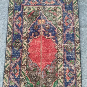 May include: A rectangular, vintage rug with a central red diamond shape, framed by a blue border with floral patterns. The rug features a distressed look with faded colors of green, brown, and beige, suggesting an antique or well-worn aesthetic.