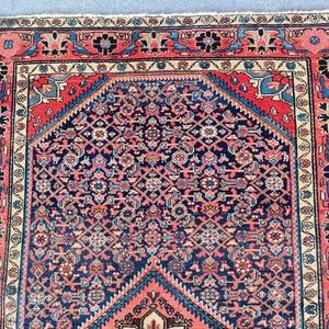 Peut inclure: Un tapis rectangulaire pr&eacute;sentant un champ central bleu fonc&eacute; avec un motif g&eacute;om&eacute;trique complexe en rouge, rose et bleu clair. Le tapis est encadr&eacute; par une large bordure aux motifs floraux rouges, bleus et cr&egrave;me, avec une finition &agrave; franges.