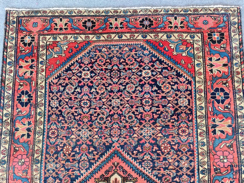 Peut inclure: Un tapis rectangulaire pr&eacute;sentant un champ central bleu fonc&eacute; avec un motif g&eacute;om&eacute;trique complexe en rouge, rose et bleu clair. Le tapis est encadr&eacute; par une large bordure aux motifs floraux rouges, bleus et cr&egrave;me, avec une finition &agrave; franges.