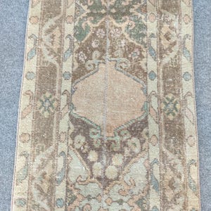 Könnte beinhalten: Ein rechteckiger Läufer mit einem zentralen Medaillon-Design in Braun-, Salbeigrün- und sanften Pfirsichtönen. Der Teppich zeigt ein symmetrisches Muster mit komplexen floralen Motiven und geometrischen Bordüren in abgenutzter Optik.