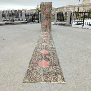 May include: A long, narrow, vintage rug with a repeating pattern of coral and brown floral medallions on a green and brown background. The rug is rolled up at one end and unrolled on a concrete surface.