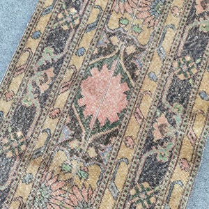 Op de afbeelding: Een rechthoekig loperkleed met een herhalend patroon van grote roze bloemmotieven in het midden van donkergrijze geometrische randen. De achtergrond is een gedempte goudbruine kleur, geaccentueerd met kleine groene en blauwe details.