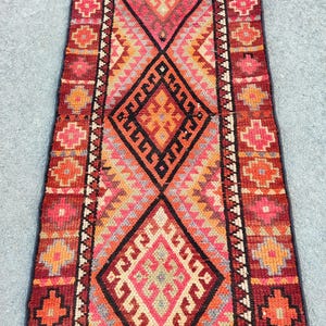 May include: A rectangular, hand-woven rug with a central diamond pattern in shades of red, orange, and pink. The rug features geometric designs and borders with floral motifs. The rug's dimensions are not visible.