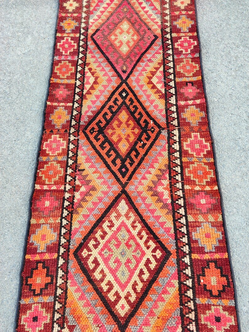 May include: A rectangular, hand-woven rug with a central diamond pattern in shades of red, orange, and pink. The rug features geometric designs and borders with floral motifs. The rug's dimensions are not visible.
