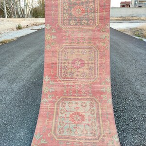 May include: A rectangular, distressed red rug with a repeating geometric pattern in beige, gray, and pink. The rug features a central medallion design and is displayed outdoors on an asphalt surface. The rug's dimensions are not visible.