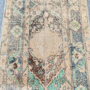 Könnte beinhalten: Ein rechteckiger Webteppich mit einem zentralen Medaillon und geometrischen Bordürenmustern in den Farben Hellbraun, Dunkelbraun und Blaugrün. Das Textil weist eine abgenutzte Struktur mit komplexen floralen und tribalartigen Motiven auf.
