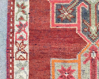 May include: Close-up of a hand-woven rug with a geometric pattern. The rug has a deep red background with accents of blue, orange, pink, and green. The design includes stylized floral and geometric shapes, creating a textured and detailed surface.