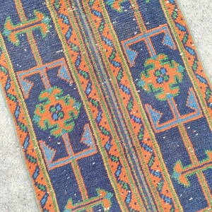 May include: A rectangular rug with a navy blue background and intricate geometric patterns in orange, green, and blue. The design features repeating stylized shapes and borders, creating a textured appearance.