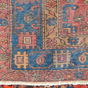 May include: Close-up of a section of a vintage rug with a detailed pattern. The rug features a blue border with geometric shapes and a central field in shades of red, brown, and beige. The rug has a fringed edge.