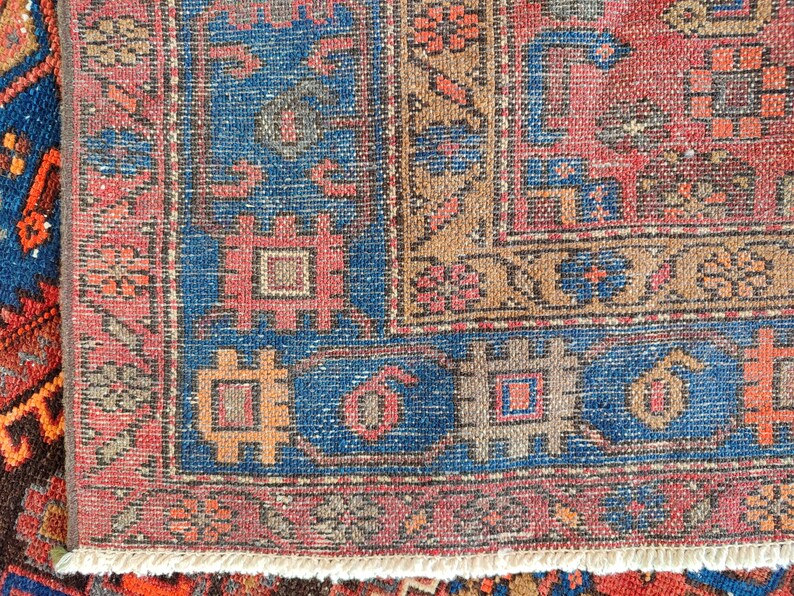 May include: Close-up of a section of a vintage rug with a detailed pattern. The rug features a blue border with geometric shapes and a central field in shades of red, brown, and beige. The rug has a fringed edge.