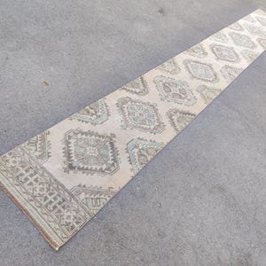 Puede incluir: Una alfombra larga y rectangular con fondo crema y patrones geométricos en tonos marrón, gris y verde azulado. La alfombra presenta un diseño de borde y motivos repetidos de diamantes y cuadrados. Mide unos 200 cm de largo.