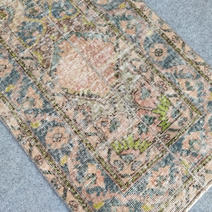 May include: A rectangular, vintage-style rug with a faded, distressed appearance. The rug features a central peach-colored field surrounded by a border of teal and olive green floral patterns. The rug's design has a classic, antique aesthetic.