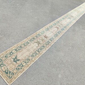 Könnte beinhalten: Ein langer, rechteckiger Teppich mit einem beigen Mittelfeld und einem Rand aus türkisfarbenen und beigen geometrischen Mustern. Der Teppich weist florale und tierische Motive auf und erzeugt eine Vintage-Ästhetik. Der Teppich liegt auf einer grauen Oberfläche.