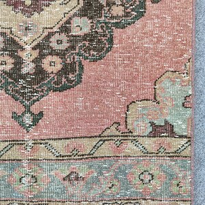 Peut inclure: Fragment de tapis vintage à l'aspect vieilli et délavé. Le tapis présente un motif central dans des tons de marron, beige et vert, sur un fond rose. Une bordure avec des motifs géométriques beiges, verts et bleu-gris encadre le motif.
