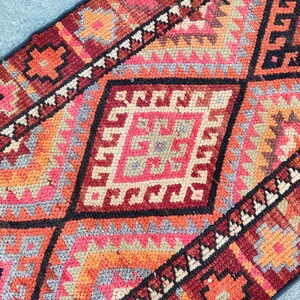 May include: Close-up of a colorful, patterned rug with geometric designs. The rug features a diamond pattern in shades of red, orange, pink, and black. The rug's design includes various shapes and patterns, creating a visually appealing textile.