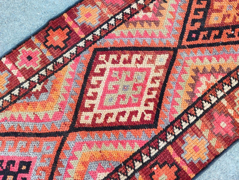 May include: Close-up of a colorful, patterned rug with geometric designs. The rug features a diamond pattern in shades of red, orange, pink, and black. The rug's design includes various shapes and patterns, creating a visually appealing textile.