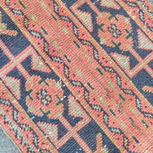 May include: Close-up of a vintage rug with a repeating geometric pattern. The rug features a combination of colours, including navy blue, coral, and olive green. The design includes floral and geometric shapes.