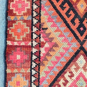 May include: Close-up of a section of a colorful, patterned rug. The rug features geometric and floral designs in shades of red, orange, pink, and black. The rug has a textured appearance.