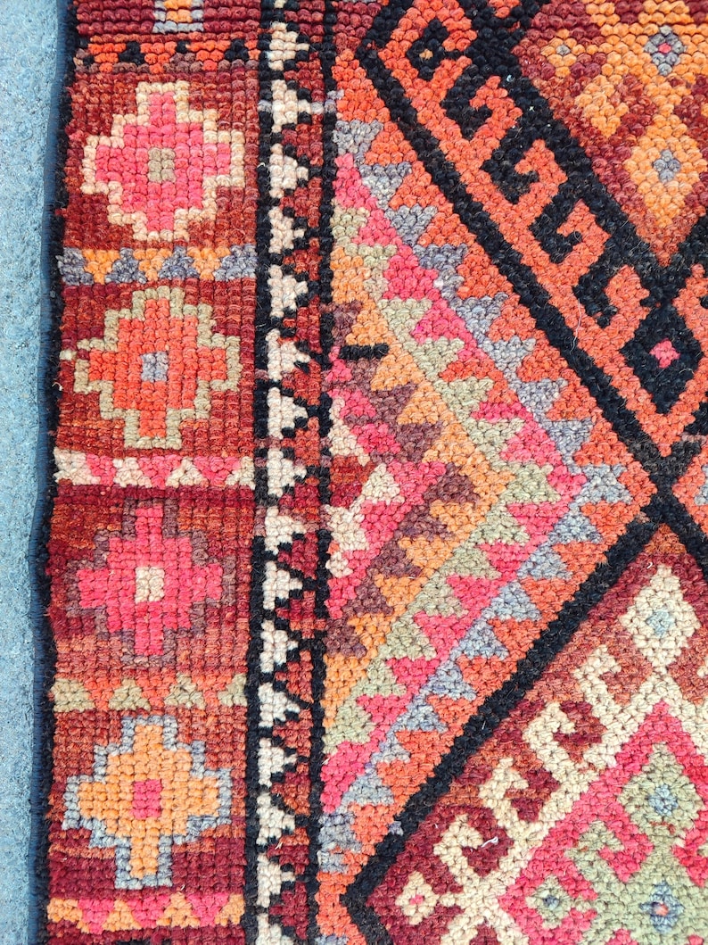 May include: Close-up of a section of a colorful, patterned rug. The rug features geometric and floral designs in shades of red, orange, pink, and black. The rug has a textured appearance.