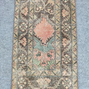Könnte beinhalten: Ein rechteckiger Vintage-Teppich mit einem zentralen Design in Pfirsich, Grün und Braun. Der Teppich hat einen Used-Look mit komplizierten floralen und geometrischen Mustern, die von einem Rand in Braun- und Beigetönen umrahmt werden. Geeignet für einen Flur oder Eingangsbereich.